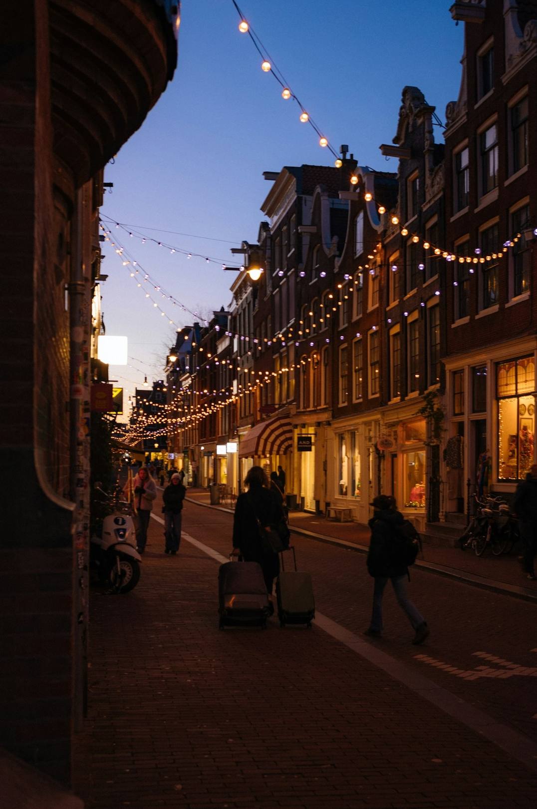People walking down a city street at dusk under string lights, unedited